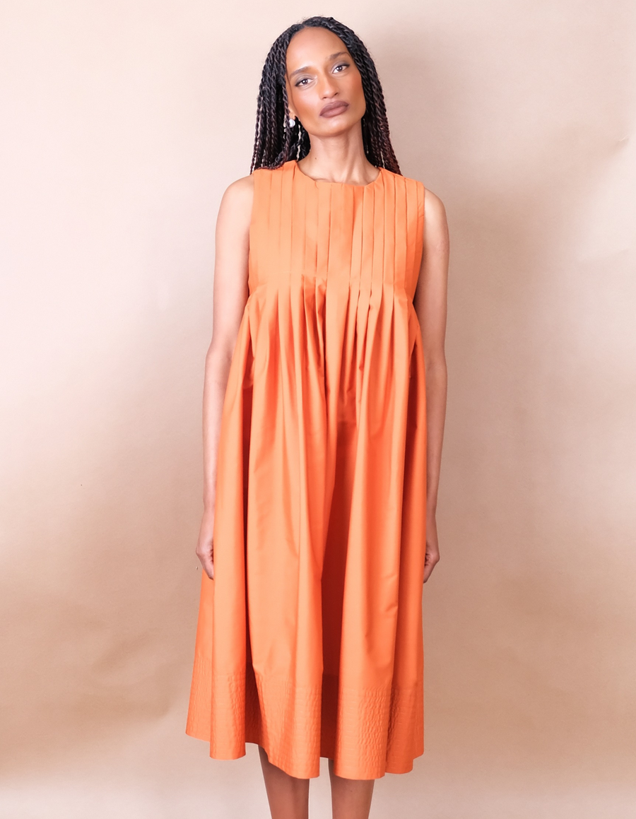 Woman wearing a orange pleated Anissa Mpungwe dress against a beige background