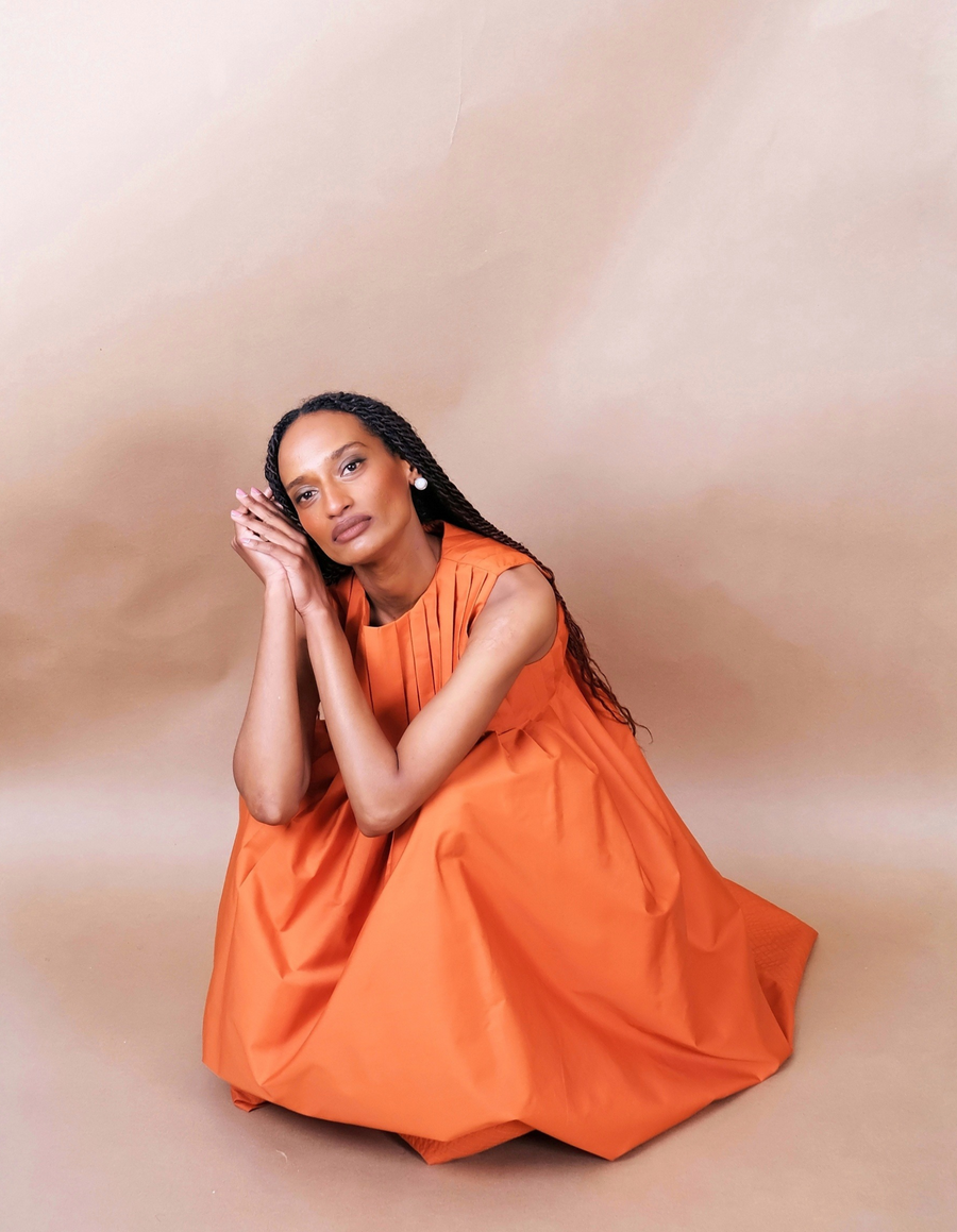 Woman wearing a orange pleated Anissa Mpungwe dress against a beige background