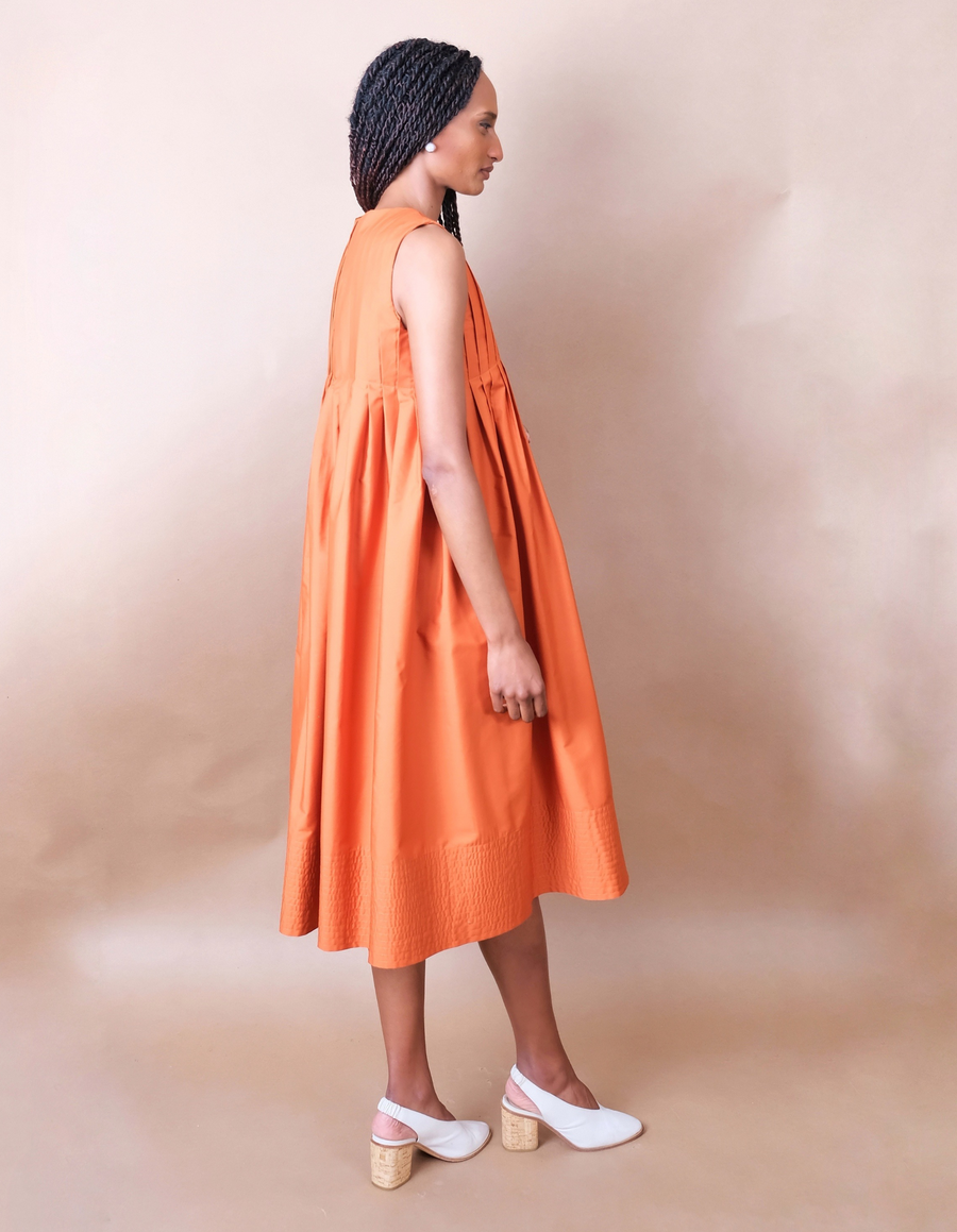 Woman wearing a orange pleated Anissa Mpungwe dress against a beige background