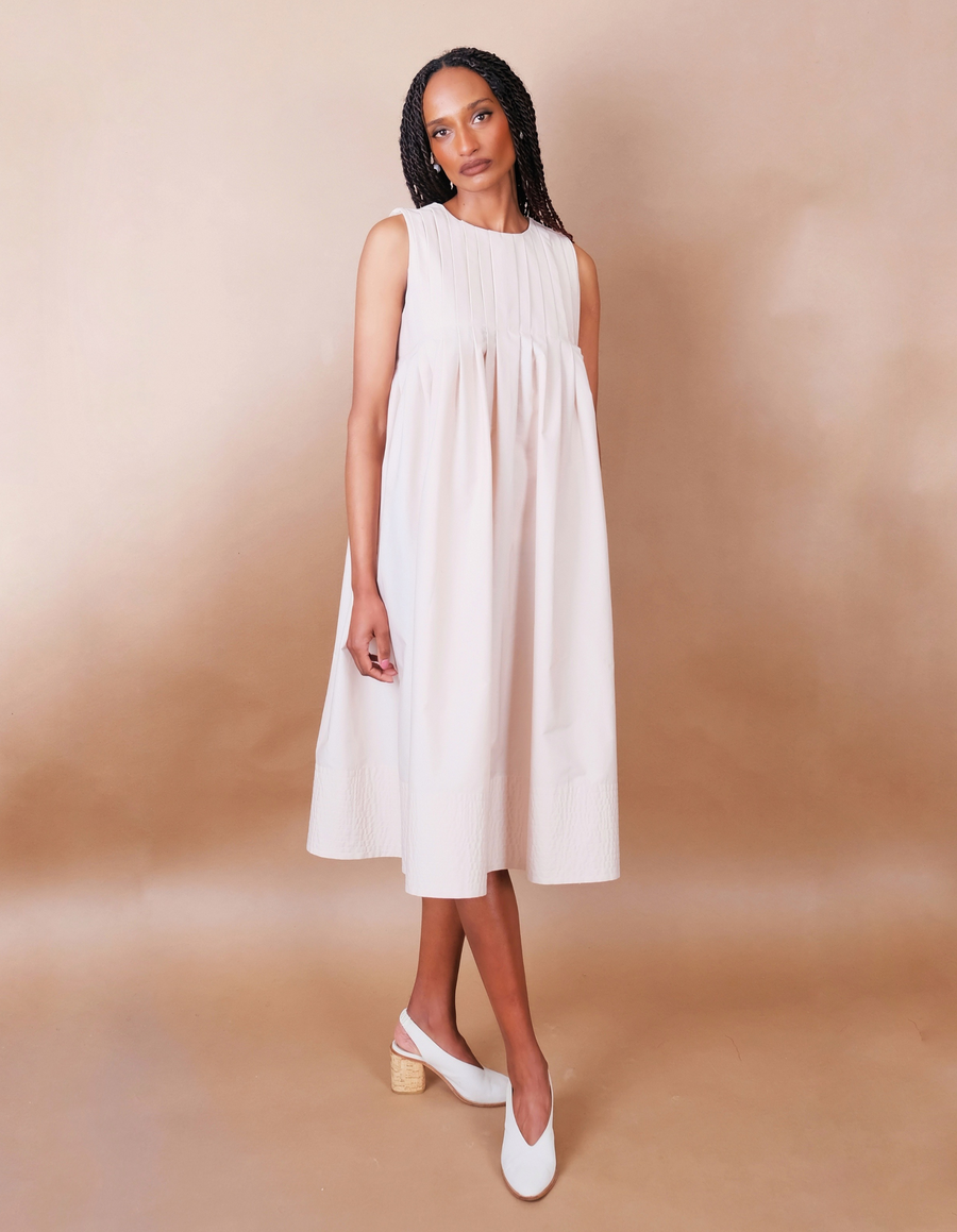 Woman wearing a white pleated Anissa Mpungwe dress against a beige background