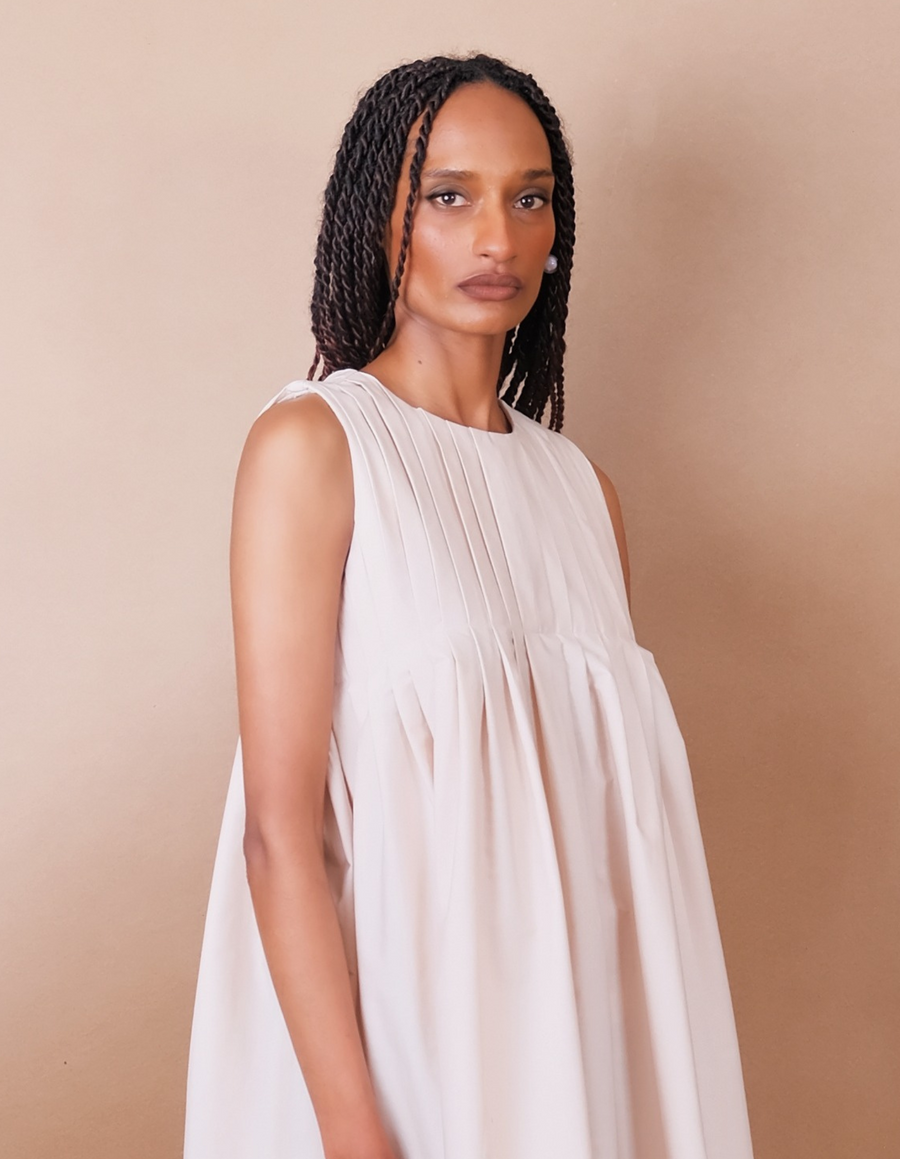 Woman wearing a white pleated Anissa Mpungwe dress against a beige background