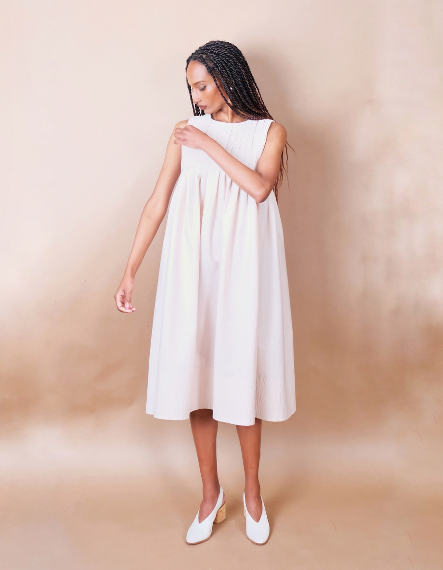 Woman wearing a white pleated Anissa Mpungwe dress against a beige background